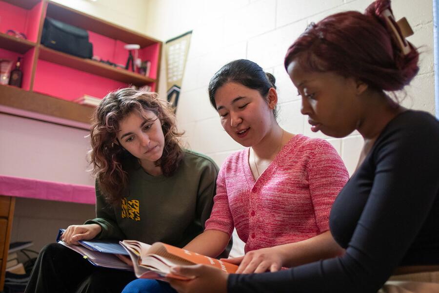 Three females looking at a book in a dorm room.